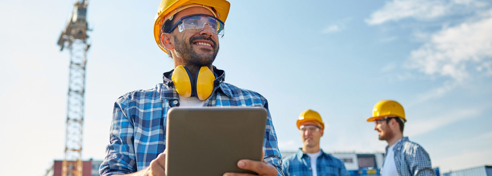 A man in a hard hat and safety glasses holding a tablet, ready to work on a construction site.